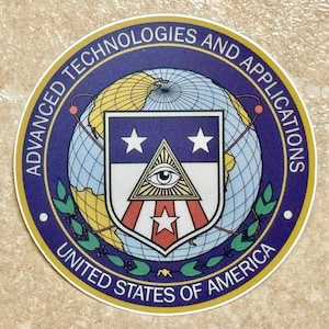 Könnte beinhalten: Ein runder Aufkleber mit blauem Rand und den Aufschriften "ADVANCED TECHNOLOGIES AND APPLICATIONS" und "UNITED STATES OF AMERICA". Das Zentrum zeigt einen Globus, ein Schild mit Sternen und ein Auge.