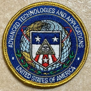 Könnte beinhalten: Runder, gestickter Aufnäher mit blau-goldenem Rand. Der Aufnäher zeigt den Text "ADVANCED TECHNOLOGIES AND APPLICATIONS" und "UNITED STATES OF AMERICA". Das zentrale Design umfasst einen Globus, Sterne und eine Pyramide mit einem Auge.