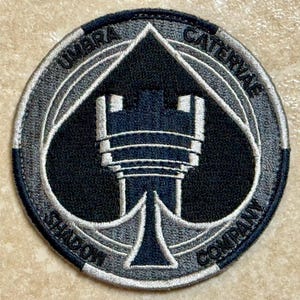 Op de afbeelding: Een ronde patch met de tekst "UMBRA CATERVAE SHADOW COMPANY" in witte letters. De patch heeft een zwarte schoppenvorm met een gestileerde toren in het midden, omlijnd in wit en grijs.