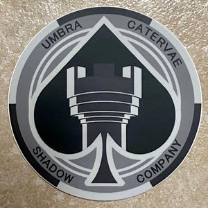 Puede incluir: Una pegatina circular con un diseño en blanco y negro. El diseño presenta una torre estilizada dentro de una forma de pica, rodeada por las palabras "UMBRA CATERVAE SHADOW COMPANY" en un patrón circular. La pegatina tiene un borde plateado.