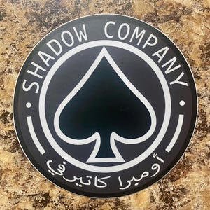 Puede incluir: Una pegatina redonda con las palabras "SHADOW COMPANY" en blanco, arqueadas en la parte superior de un círculo negro. Dentro del círculo hay un símbolo de pica negro. Texto árabe en la parte inferior.