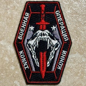 May include: A black and red embroidered patch featuring a red sword piercing a white animal skull. The patch has text in a foreign language around the edges, and two small white stars.