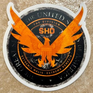 Peut inclure: Un autocollant circulaire représentant l'emblème du jeu vidéo "The Division". Le design comprend un aigle orange aux ailes déployées, le texte "SHD", et les mots "The United States of America" et "Extremis Malis Extrema Remedia" en cercle.