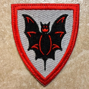 May include: A shield-shaped patch with a red border and a gray center. The patch features a black bat design with red accents. The bat has outstretched wings and a detailed body. The patch is likely an emblem or decorative item.