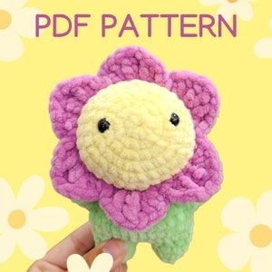 May include: A crocheted flower plush toy with a yellow face, pink petals, and green stem. The toy is being held by a hand. The background is yellow with white daisies.