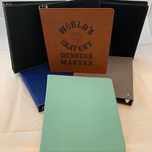 May include: A collection of six brown, black, blue, and green binders with a brown binder featuring the text "WORLD'S OKAYEST DUNGEON MASTER" with a black and white illustration of a 20-sided die.