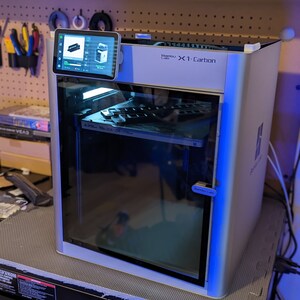 May include: A white and gray Bambu Lab X1-Carbon 3D printer with a touchscreen display. The printer has a clear front door and is sitting on a black surface. The display shows a 3D model.