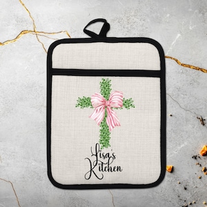 Puede incluir: Manopla de cocina color crema con borde negro y lazo para colgar. El diseño presenta una cruz verde adornada con un lazo rosa. Las palabras "Lisa's Kitchen" están escritas en una fuente de guion debajo de la cruz.
