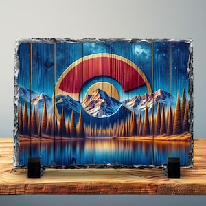 Op de afbeelding: Een rechthoekige leisteenplaat met een ontwerp van de vlag van Colorado. Het kunstwerk toont een berglandschap met een meer, bomen en het embleem van de staat in rood, goud en wit tegen een blauwe houtnerf achtergrond. De plaat wordt ondersteund door twee zwarte standaards.