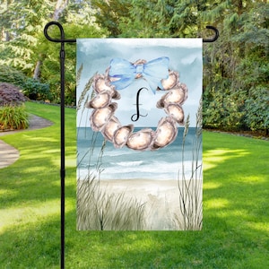 Peut inclure: Un drapeau de jardin avec un motif aquarelle d'une couronne de coquillages, un nœud bleu et la lettre "f". Le fond représente une scène de plage avec des vagues et des herbes hautes. Le drapeau est affiché sur un support en métal noir.