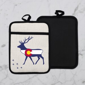 Op de afbeelding: Twee pannenlappen. Eén heeft een beige achtergrond met een zwarte rand en een Colorado-vlagontwerp in een silhouet van een hert. De andere pannenlap is effen zwart.