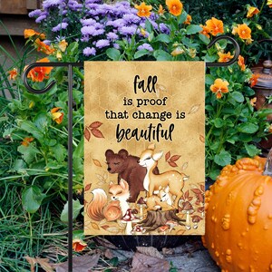 以下が含まれることがあります： 「Fall is proof that change is beautiful」のテキストと、クマ、シカ、キツネのイラストが描かれたガーデンフラッグ。秋の葉やキノコが描かれた茶色の背景です。庭に飾られています。