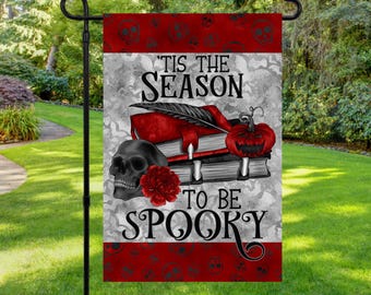 Pode incluir: Uma bandeira de jardim de Halloween com uma borda com estampa de caveiras vermelhas e pretas. A bandeira apresenta o texto "'Tis the Season to be Spooky" com uma caveira, livros, uma abóbora e uma flor vermelha.