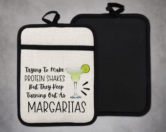 Può includere: Un sottopentola in cotone bianco con bordatura nera e il testo "Trying to make protein shakes but they keep turning out as margaritas" con un'illustrazione di margarita al lime.