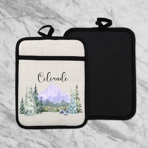 May include: Two pot holders. One features a watercolour design of a mountain range and trees with the word "Colorado" in script. The other is solid black. Both have a black loop for hanging.