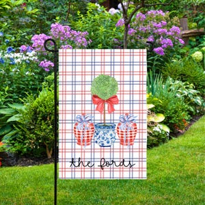 May include: A garden flag with a watercolour design of a green topiary, red bow, and two gift boxes. The flag has a red and blue plaid background, with the text "the fords" at the bottom. The flag is displayed in a garden with green grass and flowers.
