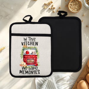 Puede incluir: Dos agarraderas. Una es negra, la otra es blanquecina con un borde negro y un lazo para colgar. La agarradera blanquecina tiene un gráfico de horno rojo con el texto "IN THIS KITCHEN We bake MEMORIES."