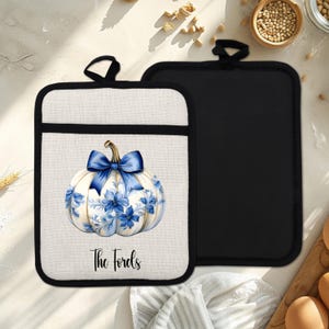 May include: Cream-coloured pot holder with a black border, featuring a blue bow and floral pumpkin design. The text "The Fords" is printed below the pumpkin. A solid black pot holder is next to it.