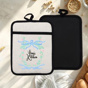 Könnte beinhalten: Zwei Topflappen. Einer ist cremefarben mit schwarzem Rand und einem Blumendesign mit dem Text "Lisa's Kitchen". Der andere ist einfarbig schwarz. Beide haben eine schwarze Schlaufe zum Aufhängen.