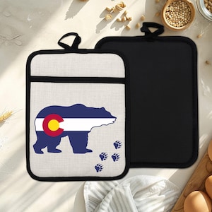 Op de afbeelding: Twee pannenlappen. Eén pannenlap heeft een berenontwerp in de kleuren van de vlag van Colorado: blauw, wit en geel. De andere is effen zwart. Beide hebben een zwarte lus om op te hangen. Afmetingen: ongeveer 20 cm x 20 cm.