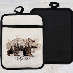 May include: Two rectangular pot holders. One is off-white with a black border and a watercolor-style grizzly bear illustration and the text "Wild One". The other is solid black.