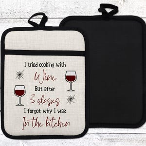 May include: A white cotton pot holder with black trim features a humorous saying about cooking with wine. The text reads "I tried cooking with wine but after 3 glasses I forgot why I was in the kitchen". The pot holder is paired with a black heat-resistant pot holder.