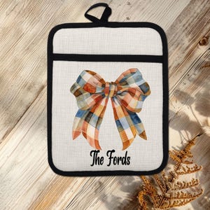 May include: An off-white rectangular pot holder with a black trim and hanging loop. It features a watercolour plaid bow in orange, blue, and brown. The text "The Fords" is printed below the bow, suitable for kitchen use.