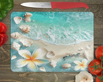 May include: A blue and white glass cutting board with a beach scene featuring seashells, waves, and a pair of sunglasses. The board is decorated with a white plumeria flower.