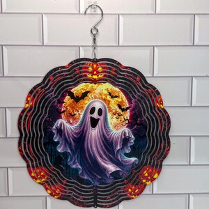 May include: A circular metal wind spinner featuring a central graphic of a white ghost against a large yellow full moon, surrounded by black bats, orange jack-o'-lanterns, and a dark purple night sky, suspended from a silver metal hook.