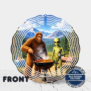 May include: A round ornament featuring a Bigfoot grilling with an alien, with a mountain backdrop. The ornament has the word "FRONT" and the text "UNIQUE HANDMADE GIFTS FOR ALL AND ALL OCCASIONS" with the logo "WILD OUTDOOR CREATIONS".