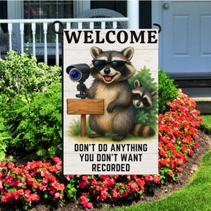 May include: A garden flag featuring a cartoon raccoon wearing sunglasses and holding a security camera. The flag reads "WELCOME" at the top and "DON'T DO ANYTHING YOU DON'T WANT RECORDED" at the bottom. The flag is displayed in a garden setting.