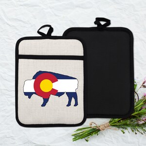 May include: Two pot holders. One has a beige background with a black border and a bison silhouette filled with the Colorado state flag. The other is solid black with a loop for hanging.