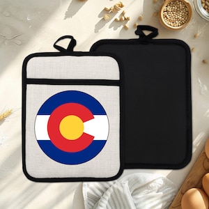 Op de afbeelding: Twee pannenlappen. Eén heeft de vlag van de staat Colorado in een cirkelvormig ontwerp met een wit, rood, blauw en geel kleurenschema. De andere is effen zwart. Beide hebben een zwarte lus om op te hangen.
