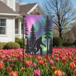 May include: A garden flag featuring a silhouette of Bigfoot walking in front of a forest scene with an aurora borealis background in shades of purple, green, and pink. The flag is displayed in a garden of colorful tulips.