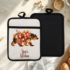 Può includere: Due presine. Una è nera, l'altra è bianco sporco con un bordo nero. La presina bianco sporco ha un disegno di orso ad acquerello con fiori e un fiocco. Il testo "Lisa's Kitchen" è sotto.