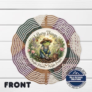 May include: A decorative garden wind spinner featuring a whimsical frog in overalls and a straw hat, digging in a garden. The spinner has a circular design with text that reads "I Bury Things... Maybe Plants... Maybe People."