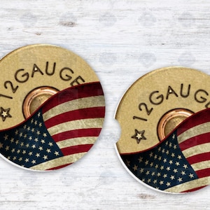 May include: Two round 12 gauge shotgun shell coasters with an American flag design. The coasters are made of a cork-like material and have a brown background with a gold rim.