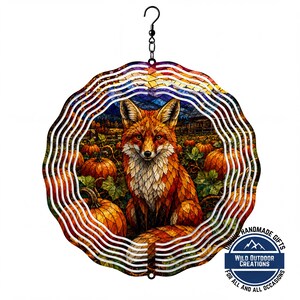 May include: A circular metal wind spinner featuring a stained-glass style illustration of a fox sitting in a pumpkin patch. The design includes orange pumpkins, green leaves, and a dark blue sky. A black swivel hook is attached to the top for hanging.