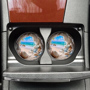 May include: Two round car coasters with a beach scene. The coasters feature a hole in the center and a beach scene with a blue ocean, white sand, and seashells.