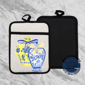 May include: Two pot holders. One features a yellow and blue vase design with a bow, the other is solid black. Both have a black loop for hanging. The design is on a beige background with a black border.