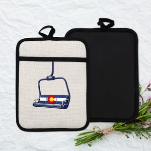 May include: Two pot holders. One is off-white with a black border and a blue ski lift graphic with the Colorado state flag. The other is solid black. Both have a black loop for hanging.
