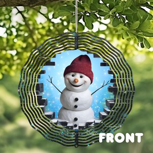 May include: A round, layered ornament featuring a smiling snowman wearing a burgundy hat and twig arms. The snowman is set against a blue, snowy background, framed by a decorative, wavy border. The word "FRONT" is printed at the bottom.
