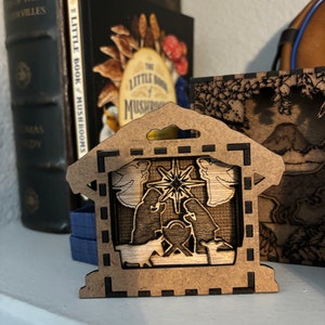May include: A wooden Christmas ornament depicting the nativity scene. The ornament is brown with a black border and features a cutout of the manger, Mary, Joseph, and the baby Jesus. There are also two angels and a star above the manger.