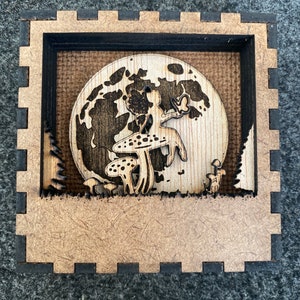 May include: A wooden box with a cutout design of a fairy standing on a mushroom in front of a full moon. The box is made of wood and has a rustic brown finish.