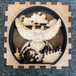 May include: A wooden owl with outstretched wings flying over a full moon and a forest. The owl is in front of a cloud and the moon is behind a forest. The scene is framed in a wooden square.