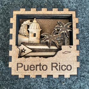 May include: A wooden cutout of a fort with palm trees and the Puerto Rican flag. The cutout is framed in a wooden square with a black border and the words "Puerto Rico" are engraved below the cutout.
