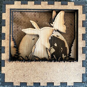 May include: A wooden shadow box with a laser-cut design of a rabbit, butterfly, and a figure silhouette in a natural scene. The box has a light brown frame with a dark brown border and a textured backdrop.