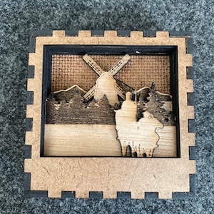 May include: A wooden shadow box with a 3D laser cut design of a windmill, trees, and hills. The background is a brown burlap fabric.
