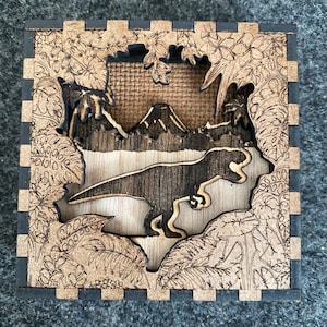 May include: A wooden box featuring a laser-cut design of a dinosaur in a jungle setting. The dinosaur is a Tyrannosaurus Rex, and the jungle is depicted with palm trees and ferns. The box is made of dark wood with a black border.