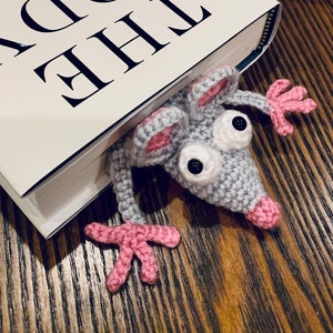 May include: A gray crocheted mouse bookmark with pink ears and paws is peeking out from under a book titled "The Body" by Bill Bryson. The book is on a wooden surface.